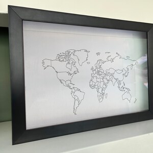 May include: A rectangular black frame containing a white print of a world map. The map features thin black outlines of continents and national borders against a plain white background, presented in a simple, clean, and minimalist style.