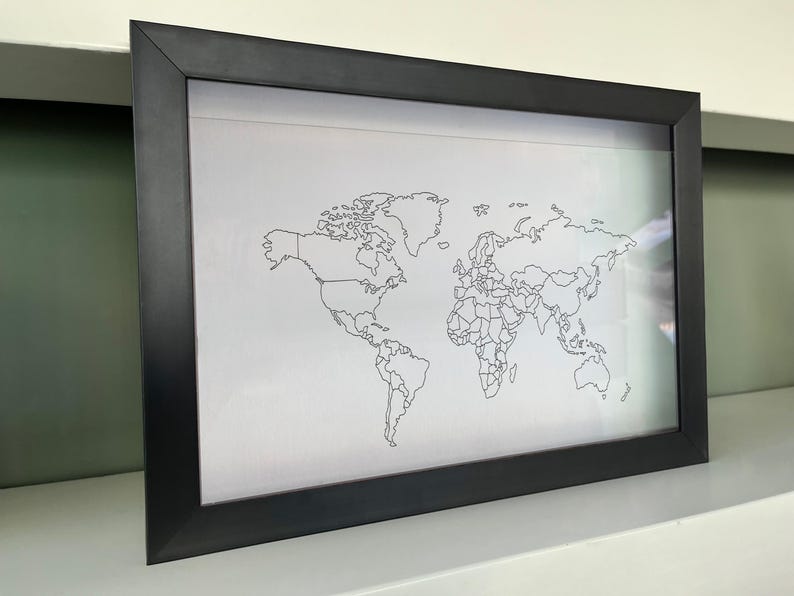 May include: A rectangular black frame containing a white print of a world map. The map features thin black outlines of continents and national borders against a plain white background, presented in a simple, clean, and minimalist style.