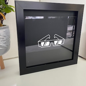 May include: A black framed square shadow box with a white outline of a pair of sunglasses inside.