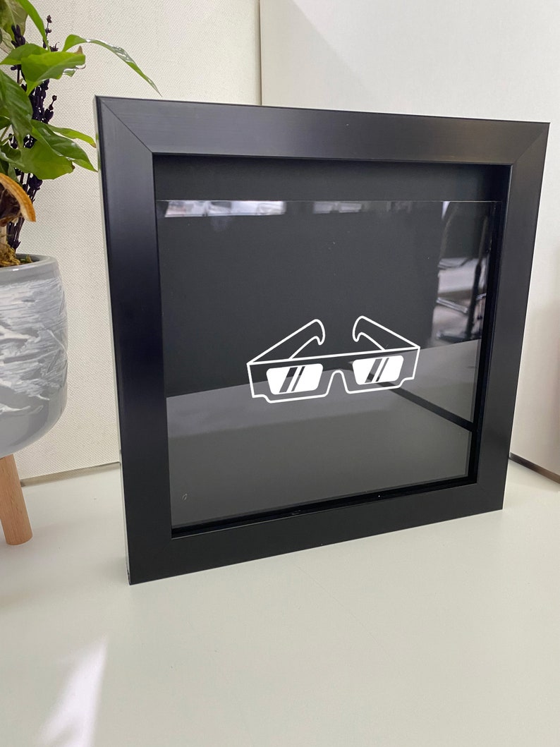 May include: A black framed square shadow box with a white outline of a pair of sunglasses inside.