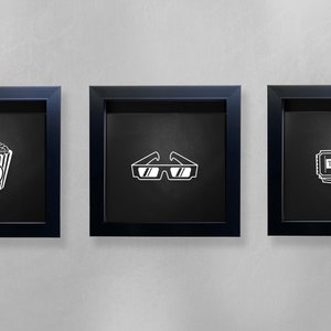 May include: Three black framed pictures on a white wall. The pictures each contain a white silhouette of a movie-related item. The first picture shows a bucket of popcorn. The second picture shows a pair of 3D glasses. The third picture shows a stack of movie tickets with the word "TICKET" printed on them.