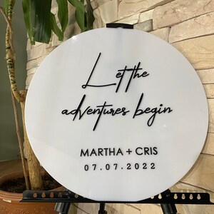 May include: White round sign with black calligraphy that reads "Let the adventures begin" and "MARTHA + CHRIS 07.07.2022".