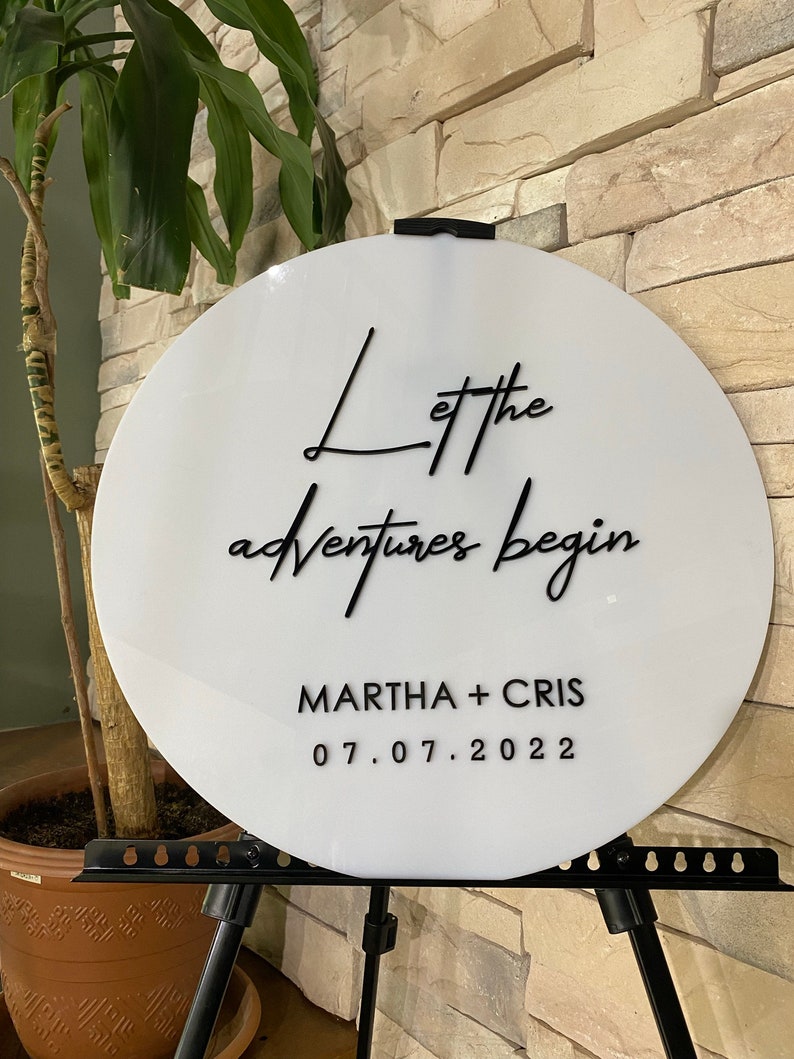 May include: White round sign with black calligraphy that reads "Let the adventures begin" and "MARTHA + CHRIS 07.07.2022".