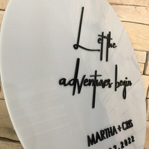 May include: White round sign with black lettering that says "Let the adventures begin" and "MARTHA + CHRIS 01.07.2022".