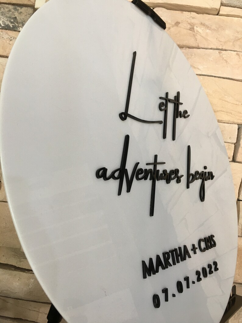 May include: White round sign with black lettering that says "Let the adventures begin" and "MARTHA + CHRIS 01.07.2022".