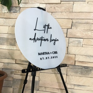 May include: White round sign with black text that reads "Let the adventures begin" and "MARTHA + CHRIS 07.01.2022". The sign is on a black easel.