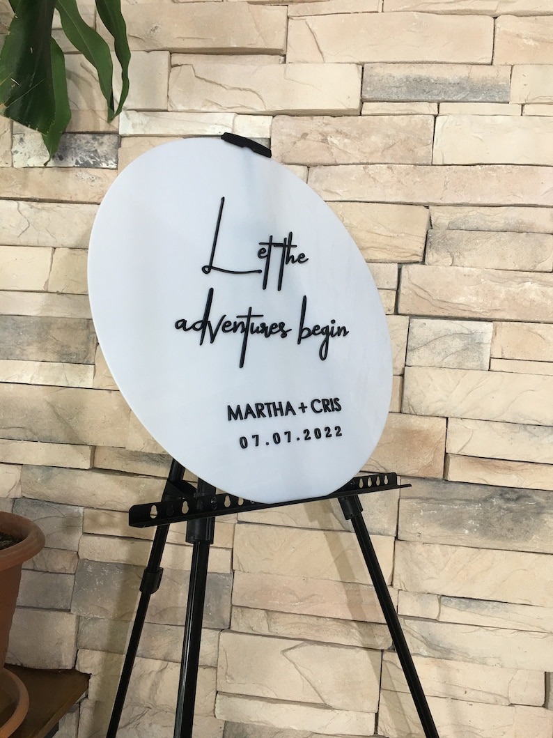 May include: White round sign with black text that reads "Let the adventures begin" and "MARTHA + CHRIS 07.01.2022". The sign is on a black easel.