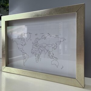 May include: A rectangular wall art piece featuring a black line drawing of a world map on a white background, encased in a metallic silver frame with a smooth, reflective finish. The map displays continental outlines and borders in a simple style.