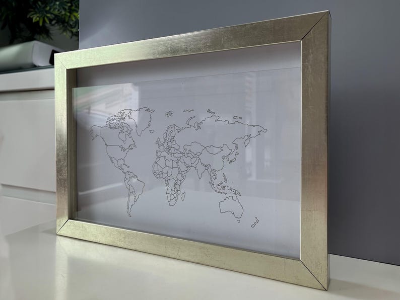 May include: A rectangular wall art piece featuring a black line drawing of a world map on a white background, encased in a metallic silver frame with a smooth, reflective finish. The map displays continental outlines and borders in a simple style.
