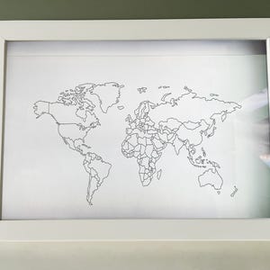 May include: A rectangular white frame containing a white print of a world map. The map features thin black outlines of continents and national borders against a plain white background, presented in a clean, simple, and minimalist style.