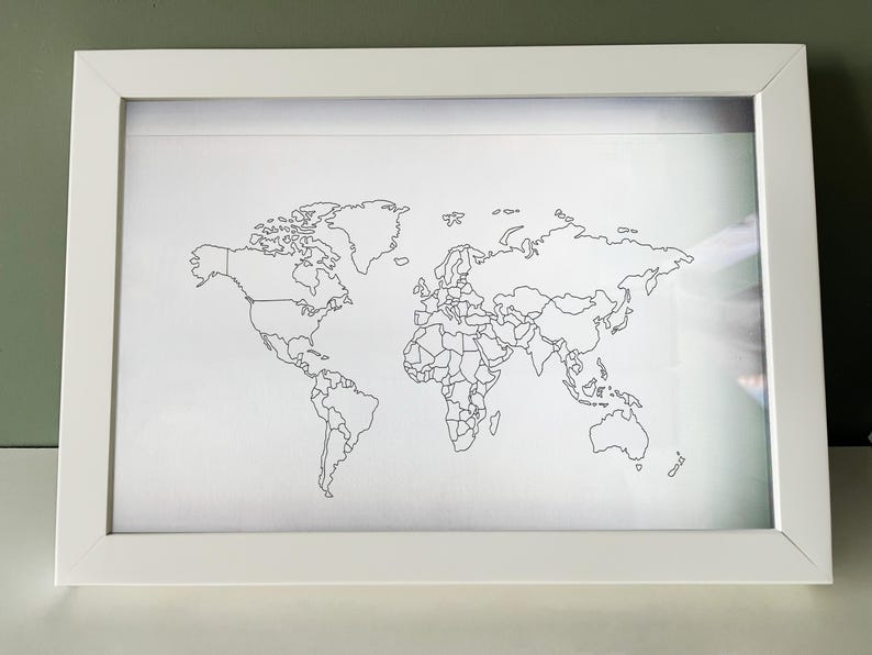 May include: A rectangular white frame containing a white print of a world map. The map features thin black outlines of continents and national borders against a plain white background, presented in a clean, simple, and minimalist style.