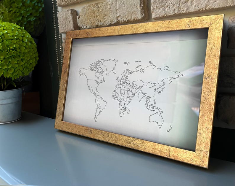 May include: A rectangular wall art piece featuring a black line drawing of a world map on a white background, encased in a textured, metallic gold frame with a distressed finish. The map displays continental outlines and country borders.