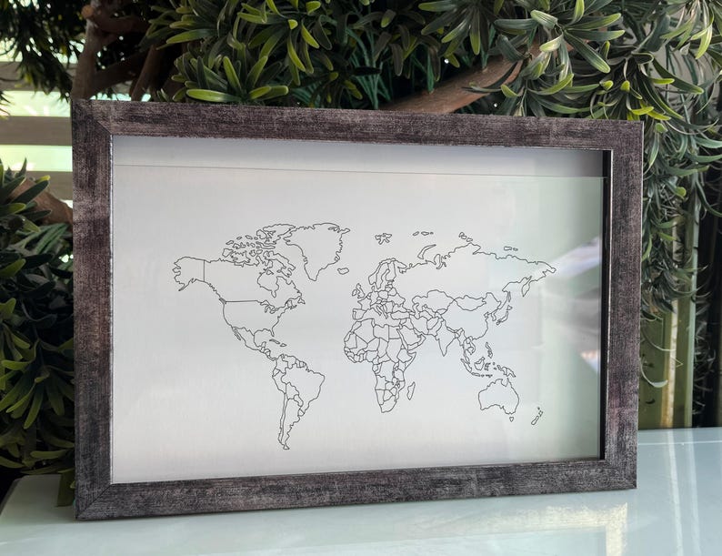 May include: A rectangular wall art piece featuring a black line drawing of a world map on a white background, encased in a dark, weathered gray wooden frame with a visible grain texture. The map displays continental outlines and borders.