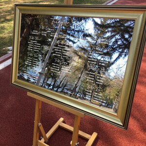 May include: A gold framed mirror with a seating chart for a wedding reception. The mirror is reflecting trees and a building in the background. The seating chart is written in white calligraphy and lists the names of guests and their assigned tables.