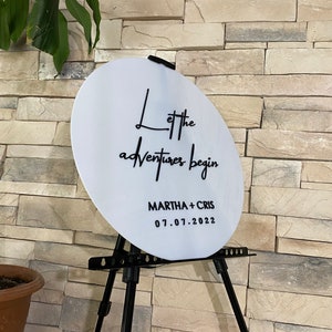 May include: White round sign with black text that reads "Let the adventures begin" and "MARTHA + CHRIS 07.01.2022". The sign is on a black easel.