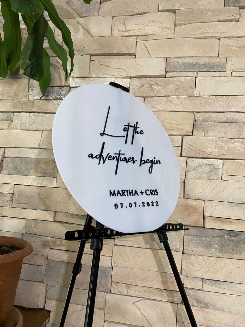 May include: White round sign with black text that reads "Let the adventures begin" and "MARTHA + CHRIS 07.01.2022". The sign is on a black easel.