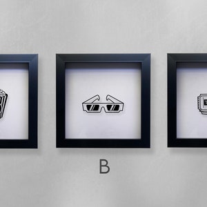 May include: Three black framed shadow boxes with white backgrounds. The shadow boxes are labeled A, B, and C. Shadow box A contains a black and white illustration of a bucket of popcorn. Shadow box B contains a black and white illustration of 3D glasses. Shadow box C contains a black and white illustration of a movie ticket.