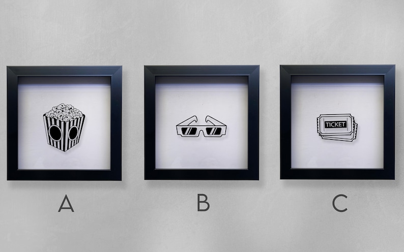 May include: Three black framed shadow boxes with white backgrounds. The shadow boxes are labeled A, B, and C. Shadow box A contains a black and white illustration of a bucket of popcorn. Shadow box B contains a black and white illustration of 3D glasses. Shadow box C contains a black and white illustration of a movie ticket.