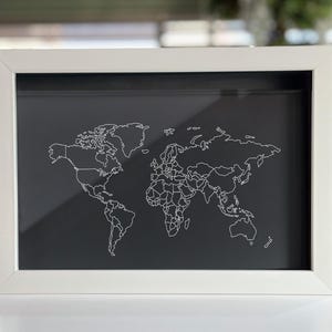 May include: A rectangular shadow box frame with a white border containing a black background. A white line drawing of a world map featuring continental outlines and country borders is displayed on the black surface within the frame.