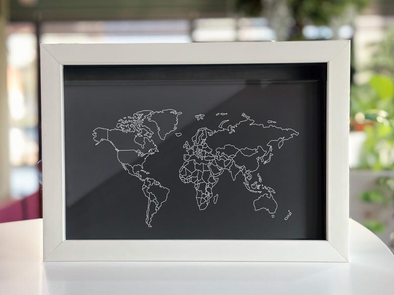 May include: A rectangular shadow box frame with a white border containing a black background. A white line drawing of a world map featuring continental outlines and country borders is displayed on the black surface within the frame.