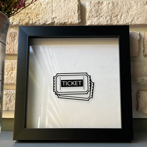 May include: A black framed shadow box with a white background and a black and white illustration of a ticket with the word "TICKET" printed on it.