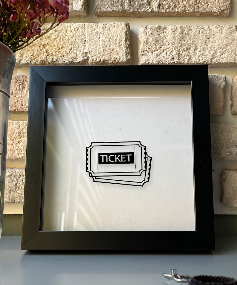 May include: A black framed shadow box with a white background and a black and white illustration of a ticket with the word "TICKET" printed on it.