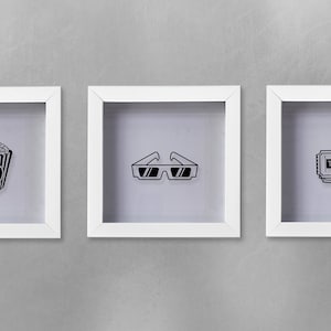 May include: Three white picture frames with black and white illustrations of movie-themed items. The frames are hanging on a gray wall. The illustrations are of a bucket of popcorn, 3D glasses, and a movie ticket.