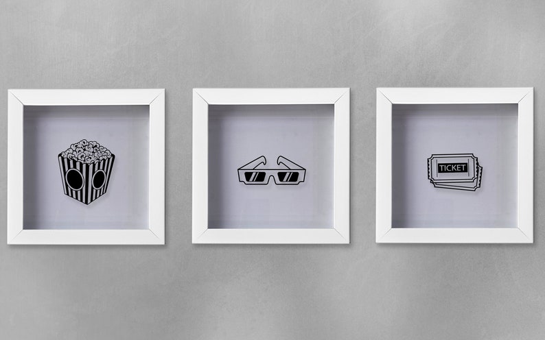 May include: Three white picture frames with black and white illustrations of movie-themed items. The frames are hanging on a gray wall. The illustrations are of a bucket of popcorn, 3D glasses, and a movie ticket.
