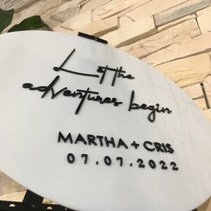 May include: White round sign with black lettering that reads "Let the adventures begin Martha + Cris 07.07.2022".