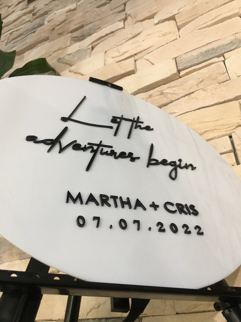May include: White round sign with black lettering that reads "Let the adventures begin Martha + Cris 07.07.2022".