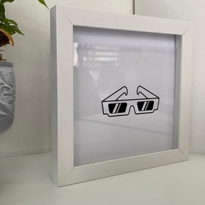 May include: A white picture frame with a clear acrylic front. The frame contains a black and white line drawing of a pair of 3D glasses.