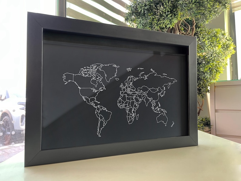 May include: A rectangular black frame containing a dark background with a white line art world map. The map features thin, continuous outlines of continents and country borders, centered within the frame, creating a high-contrast visual display.