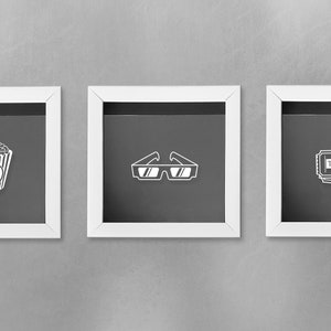 May include: Three white picture frames with black mats, each featuring a different movie-themed icon: a bucket of popcorn, 3D glasses, and a movie ticket.