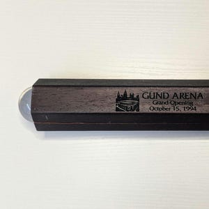 May include: A rectangular, dark wood object with a clear, rounded end. The text "GUND ARENA Grand Opening October 15, 1994" is etched on the wood, along with a graphic of a building.
