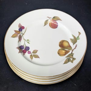 Op de afbeelding: Een stapel witte keramische borden met een gouden rand. Elk bord is versierd met een fruitontwerp, waaronder perziken, peren en bramen. De borden zijn op elkaar gestapeld.