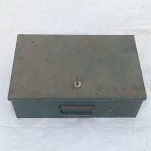 May include: A rectangular, olive-green metal box with a keyhole and a handle. The box has a slightly weathered appearance, suggesting age or use. The key is inserted in the keyhole. The box is likely used for storage.