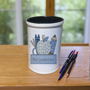 Può includere: Portapenne in ceramica bianca con bordo interno nero. Il portapenne presenta un design a mela blu con motivi floreali, pastelli, una matita e il testo "Miss Leadbetter" su un righello. Penne e una matita sono sulla superficie di legno.