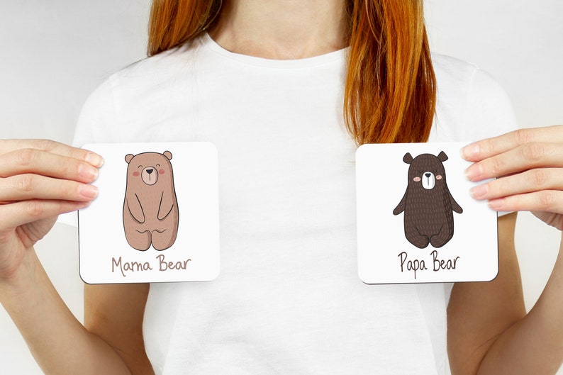 May include: Two square coasters, each featuring a cartoon bear illustration. One coaster reads "Mama Bear" and depicts a light brown bear. The other reads "Papa Bear" and shows a dark brown bear.