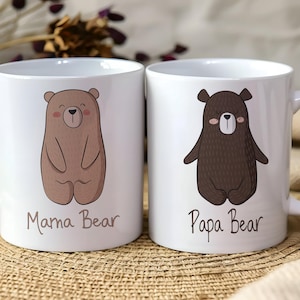 May include: Two white ceramic mugs with bear illustrations. One mug features a light brown bear with the text "Mama Bear", and the other has a dark brown bear with the text "Papa Bear". Both mugs have a curved handle.