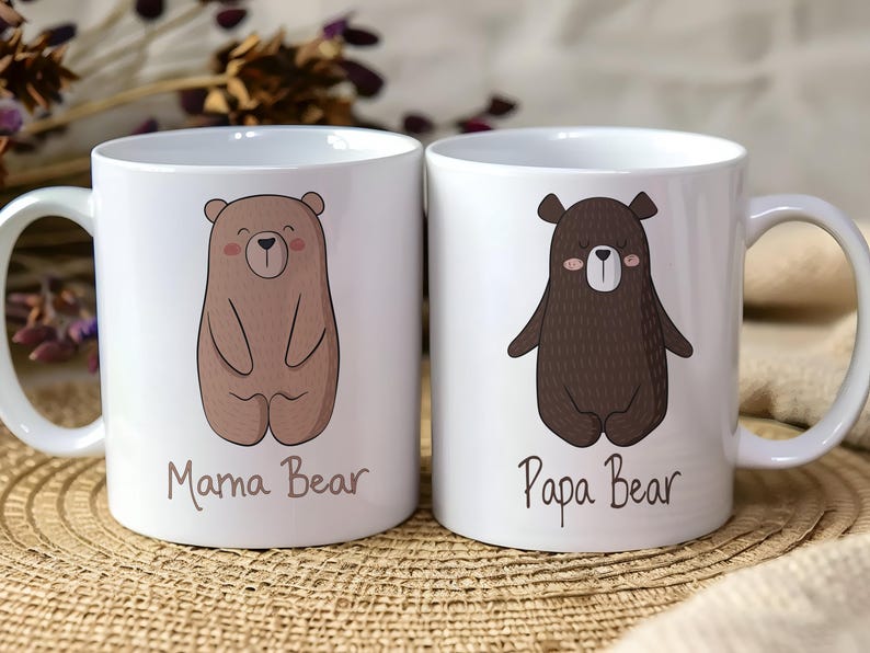 May include: Two white ceramic mugs with bear illustrations. One mug features a light brown bear with the text "Mama Bear", and the other has a dark brown bear with the text "Papa Bear". Both mugs have a curved handle.