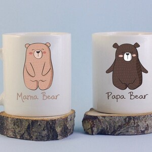 May include: Two white ceramic mugs with bear illustrations. One mug features a light brown bear with the text "Mama Bear", and the other has a dark brown bear with the text "Papa Bear". The mugs are on wooden coasters.