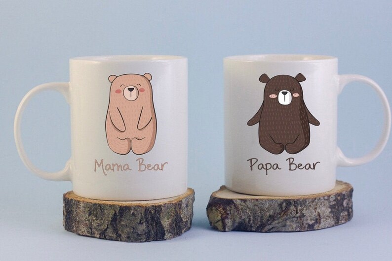 May include: Two white ceramic mugs with bear illustrations. One mug features a light brown bear with the text "Mama Bear", and the other has a dark brown bear with the text "Papa Bear". The mugs are on wooden coasters.
