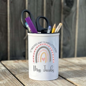 Może przedstawiać: Biały ceramiczny pojemnik na ołówki z wzorem tęczy i tekstem "Trzeba wielkiego serca, aby kształtować małe umysły". Tęcza jest ozdobiona różowymi i szarymi sercami. Tekst "Miss Jacobs" jest nadrukowany pod tęczą.