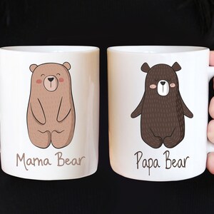 May include: Two white ceramic mugs with bear illustrations. One mug features a light brown bear with the text "Mama Bear", and the other has a dark brown bear with the text "Papa Bear". The mugs are held by a person.