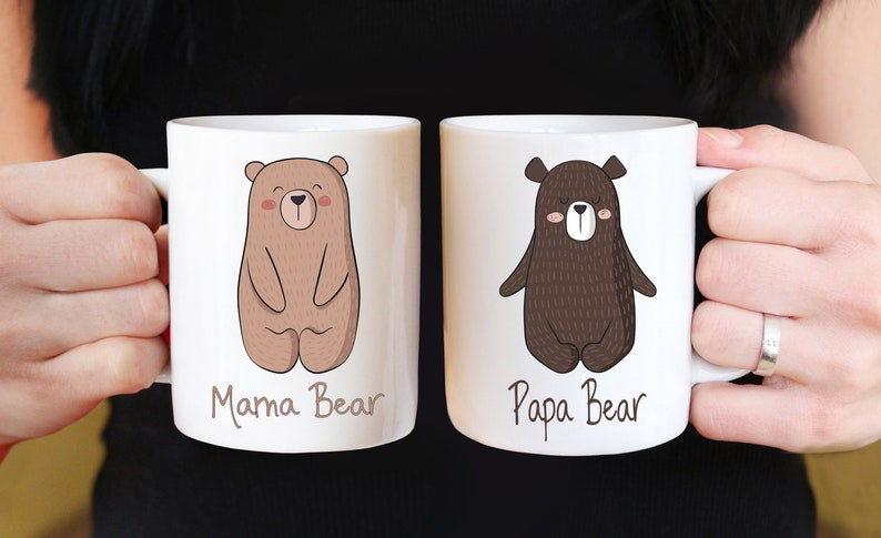 May include: Two white ceramic mugs with bear illustrations. One mug features a light brown bear with the text "Mama Bear", and the other has a dark brown bear with the text "Papa Bear". The mugs are held by a person.