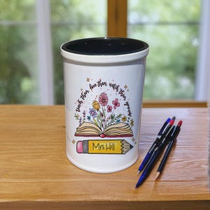 Könnte beinhalten: Weißer Keramik-Stifthalter mit schwarzem Innenraum. Der Halter zeigt ein farbenfrohes Blumendesign mit den Worten "teach them, love them, watch them grow" und einen Bleistift mit dem Namen "Mrs Hill". Stifte und ein Bleistift liegen daneben.