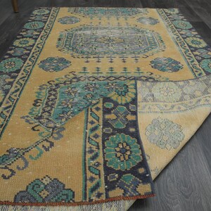 Puede incluir: Una alfombra vintage con fondo amarillo y un patrón geométrico azul y verde. La alfombra está doblada por la mitad y los bordes están deshilachados.