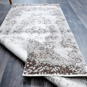 Pode incluir: Um tapete retangular com um design floral desbotado em tons de branco, cinza e marrom. O tapete está parcialmente enrolado, mostrando sua parte inferior. Ele está sobre um chão de madeira cinza. Medidas em centímetros.