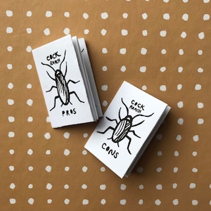 Puede incluir: Dos pequeños libros blancos con ilustraciones de cucarachas en tinta negra. Los libros se titulan "Pros de la cucaracha" y "Contras de la cucaracha".