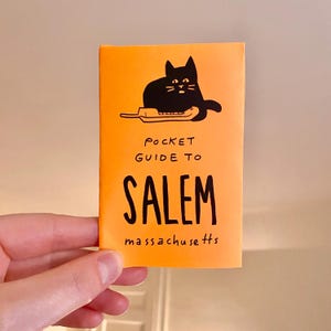 Könnte beinhalten: Orangefarbener Taschenführer mit einer schwarzen Katze, die auf einem Telefon sitzt. Der Führer trägt den Titel "Pocket Guide to Salem, Massachusetts".
